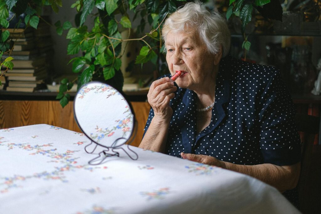 Senior woman sitting at a table applies lipstick while looking in a mirror, surrounded by plants indoors.