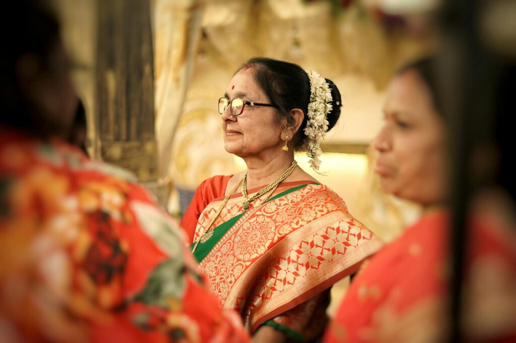 Senior woman wearing a red and gold sari, adorned with flowers, at an indoor gathering.