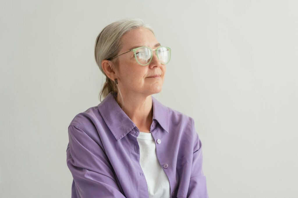 Senior woman with grey hair wearing eyeglasses and a purple shirt, looking thoughtful.
