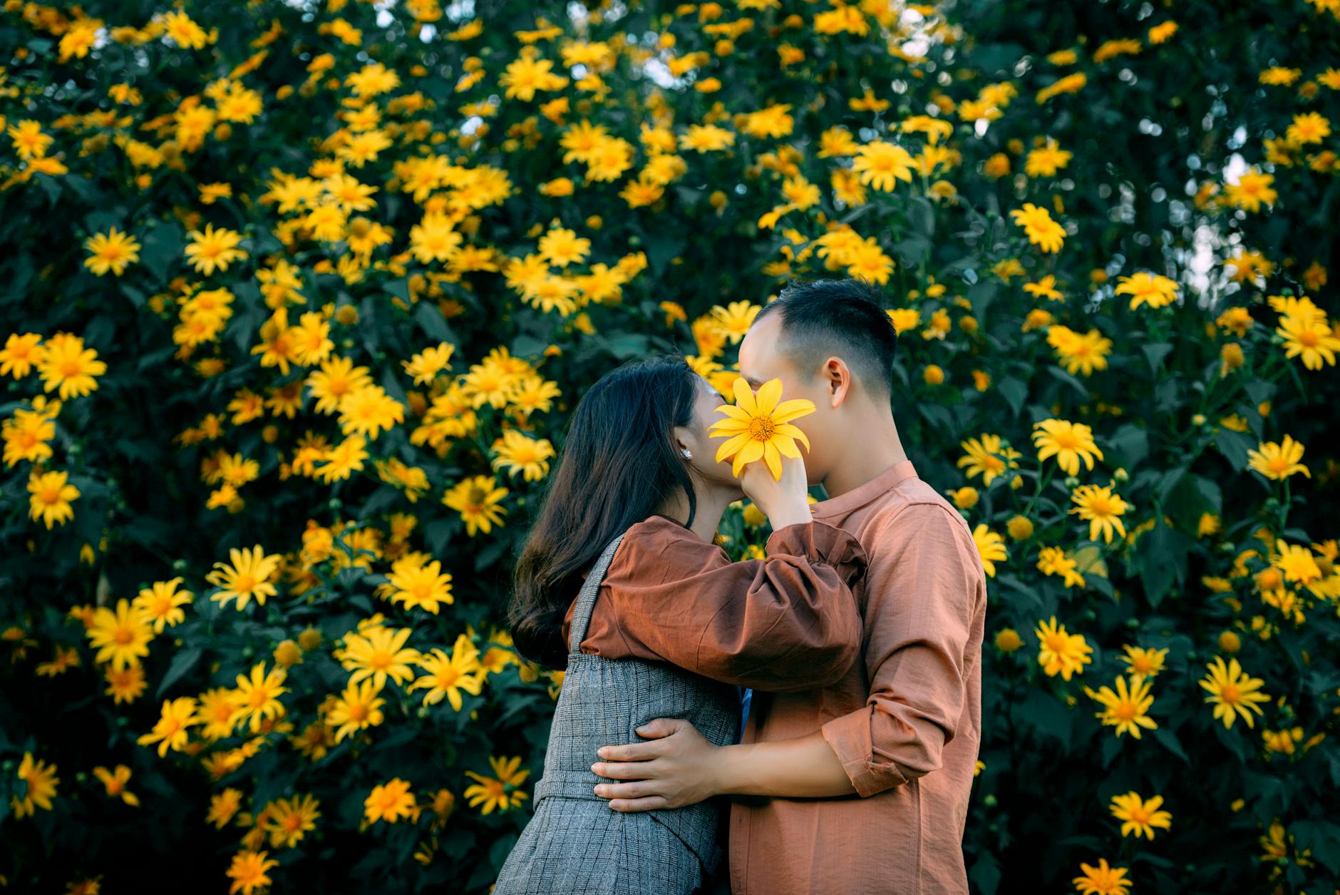 Side view of unrecognizable romantic couple embracing and covering faces with yellow flower while standing near lush blossoming bush in garden.Trung Nguyen