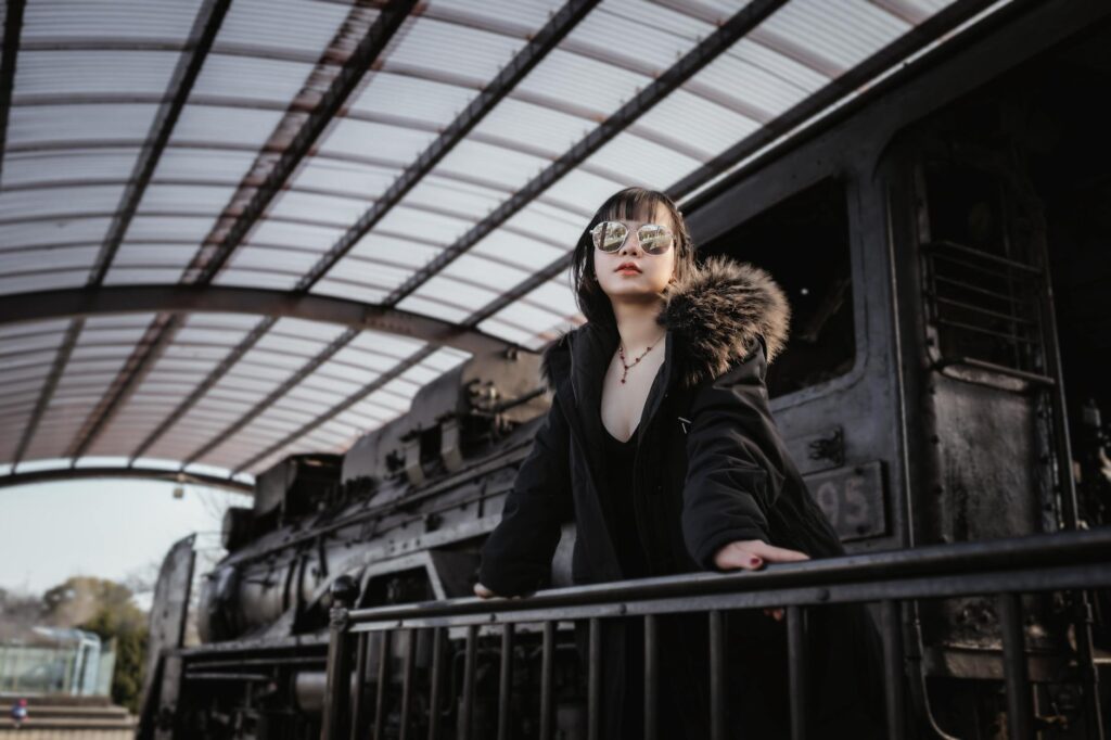Stylish woman in fur coat and sunglasses posing near an old train under a covered platform.
