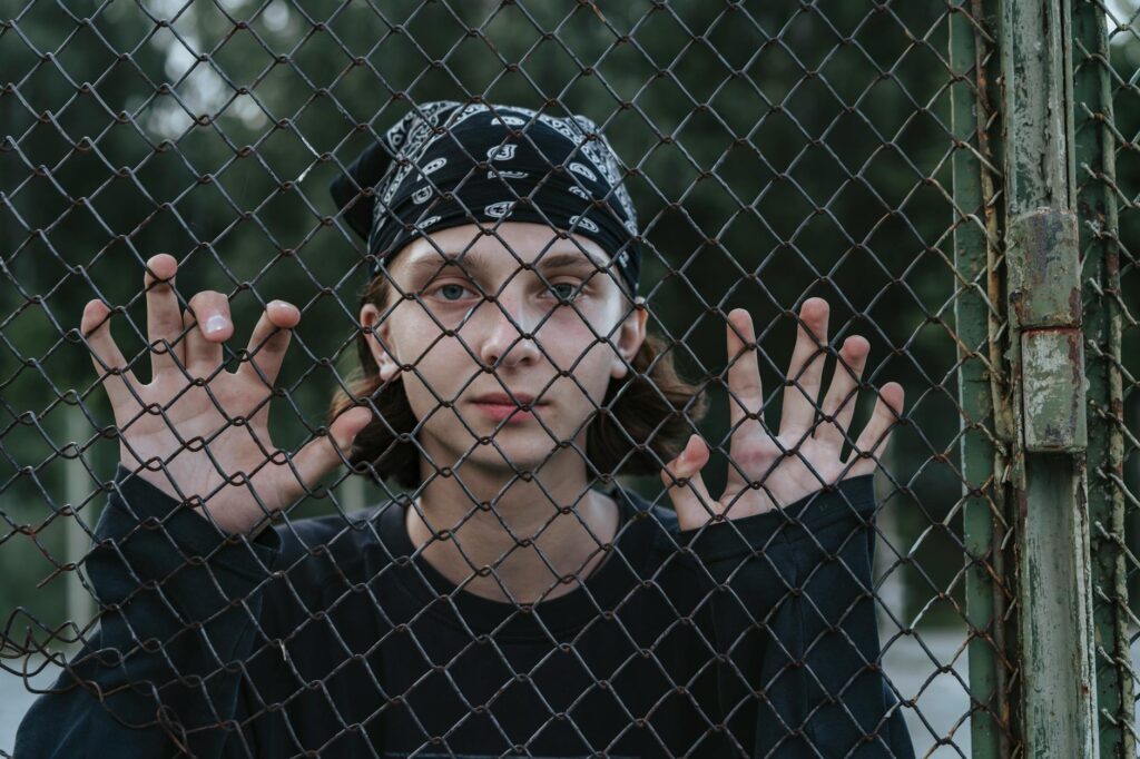 Teen wearing black bandana and long sleeves poses behind a chain link fence, symbolizing youth expression.