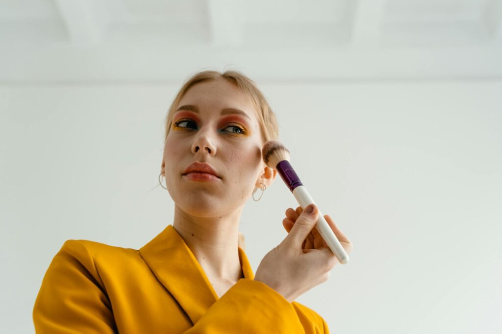 Woman in yellow attire applying blush with a makeup brush indoors.