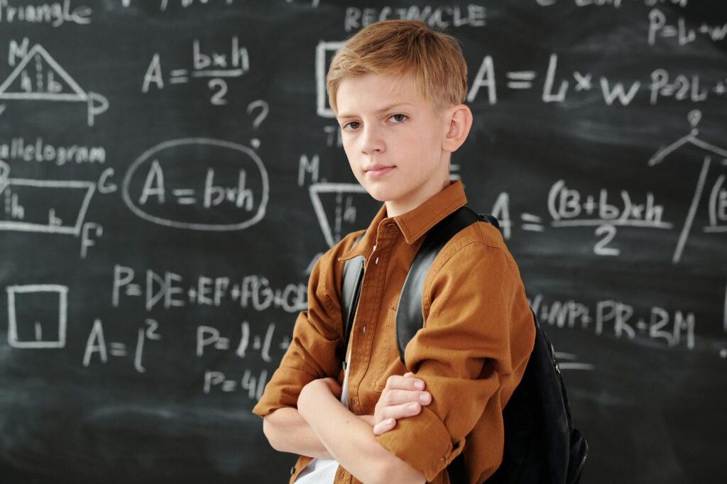 Young_boy_stands_confidently_with_arms_crossed_in_a_classroom,_in_front_of_a_blackboard_covered_with_math_equations..Max_Fischer