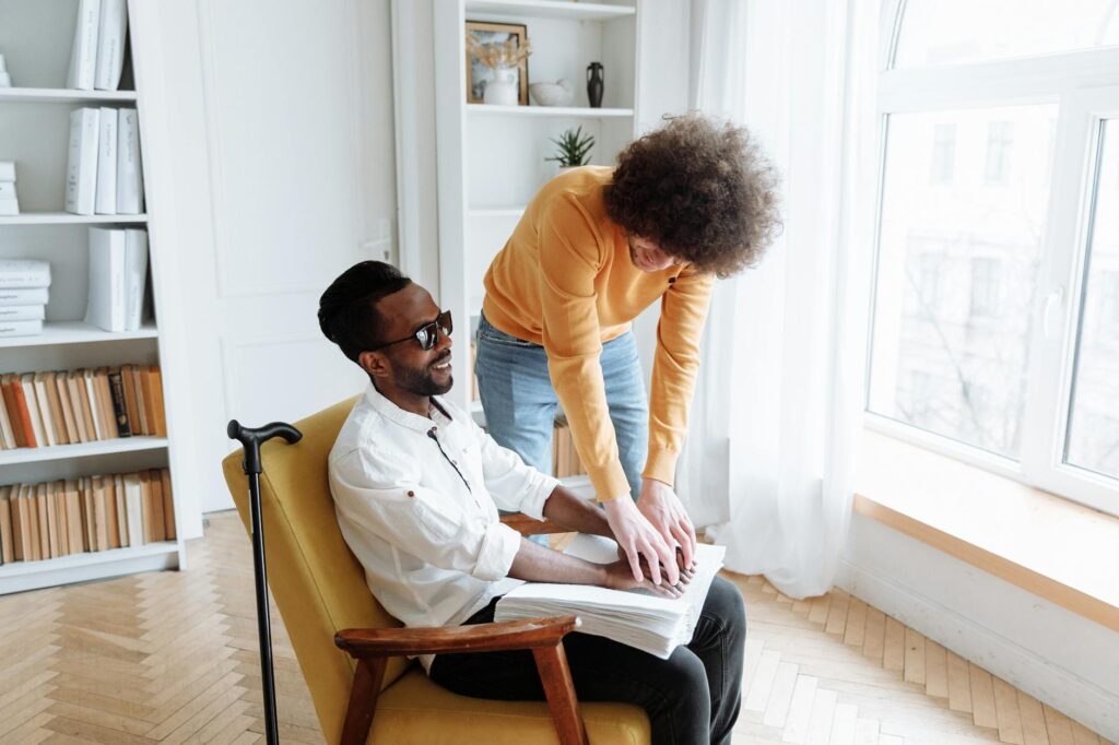Young person assists man in reading braille indoors, fostering sensory learning.