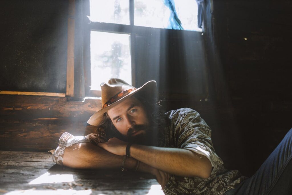 Man with a Beard and Long Hair in a Beige Hat Leaning on a Wooden Table in a Dark Room with Light from the Window