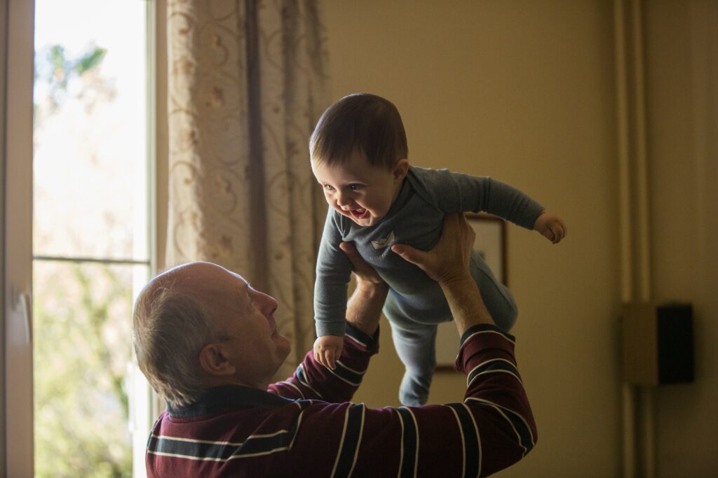 man_wearing_maroon,_white,_and_blue_stripe_long-sleeved_shirt_lifting_up_baby_wearing_gray_onesie.Johnny_Cohen