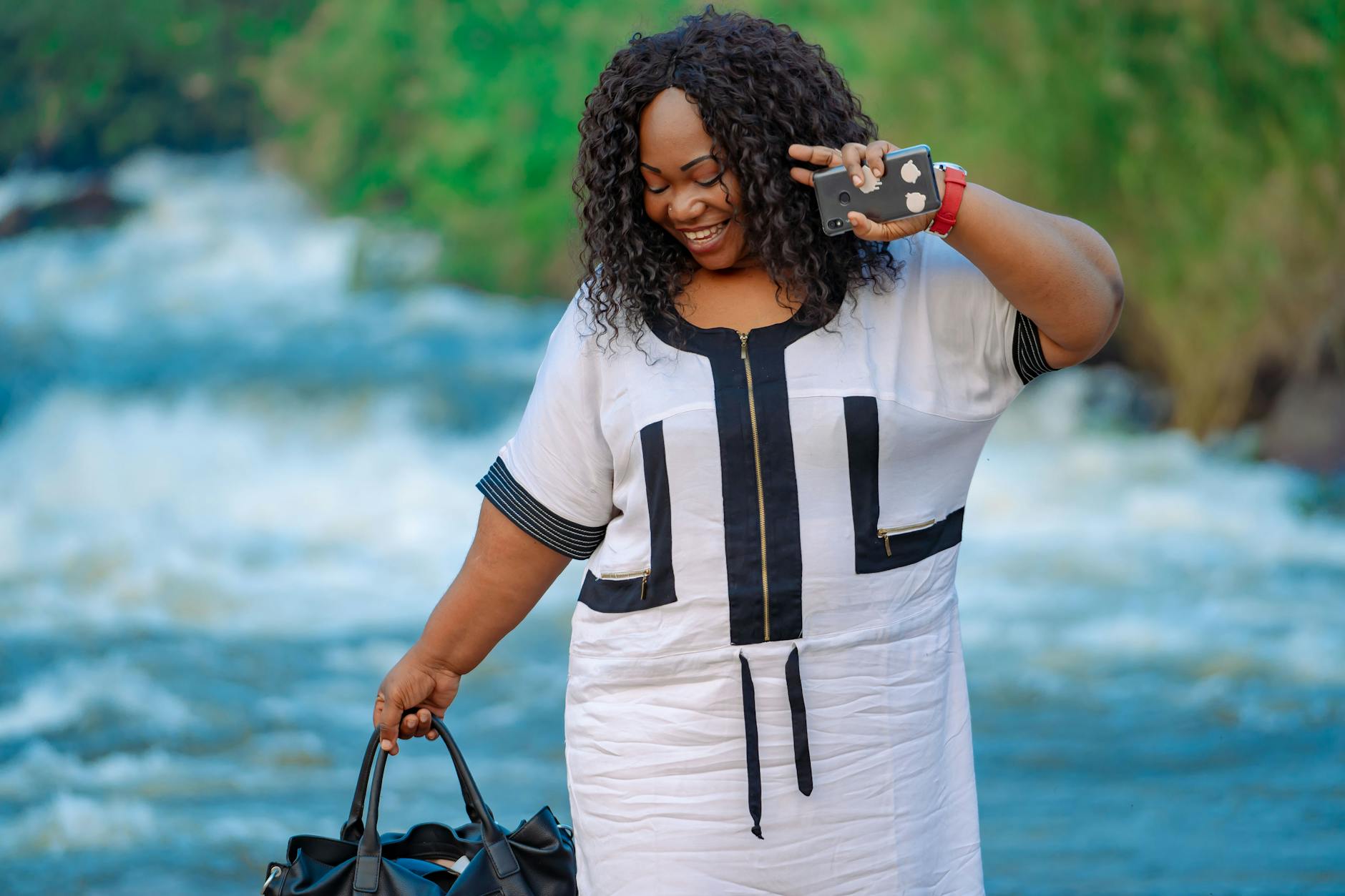 A cheerful woman in a stylish dress smiles while holding a smartphone outdoors near a scenic river.