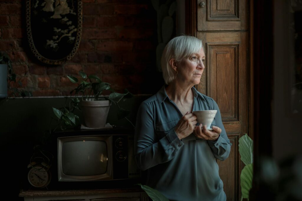 A senior woman with gray hair gazes thoughtfully out the window holding a cup, surrounded by plants.