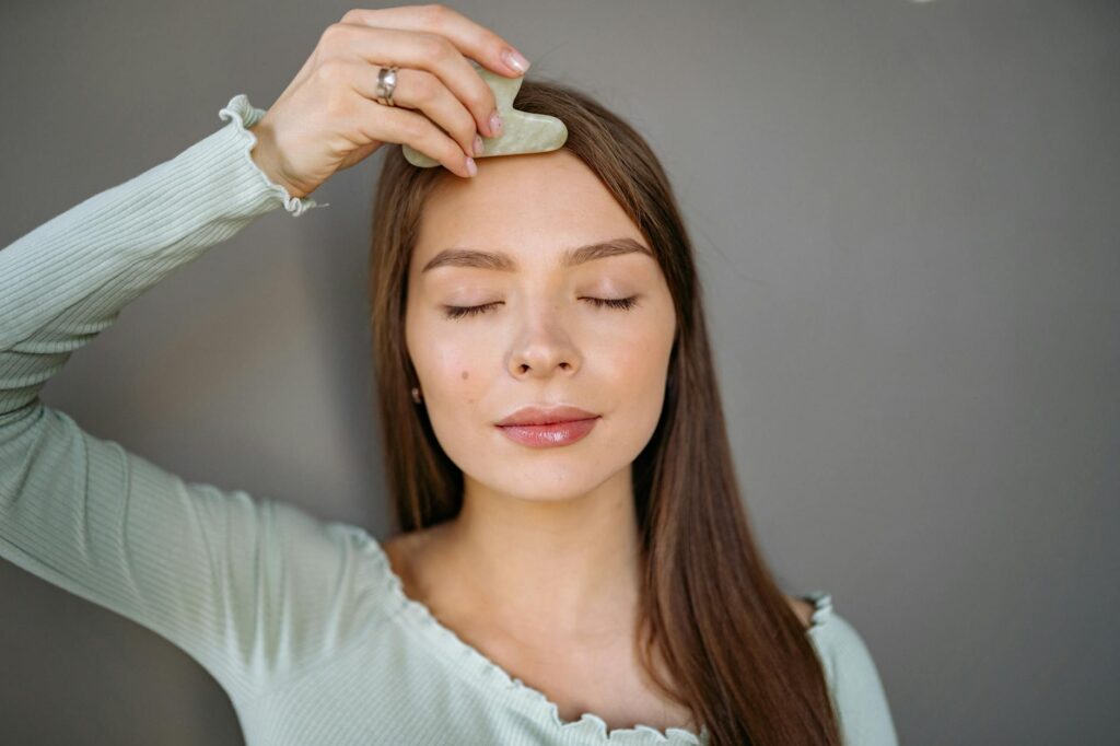 A woman relaxes with a jade gua sha tool for a facial skincare routine against a gray background.