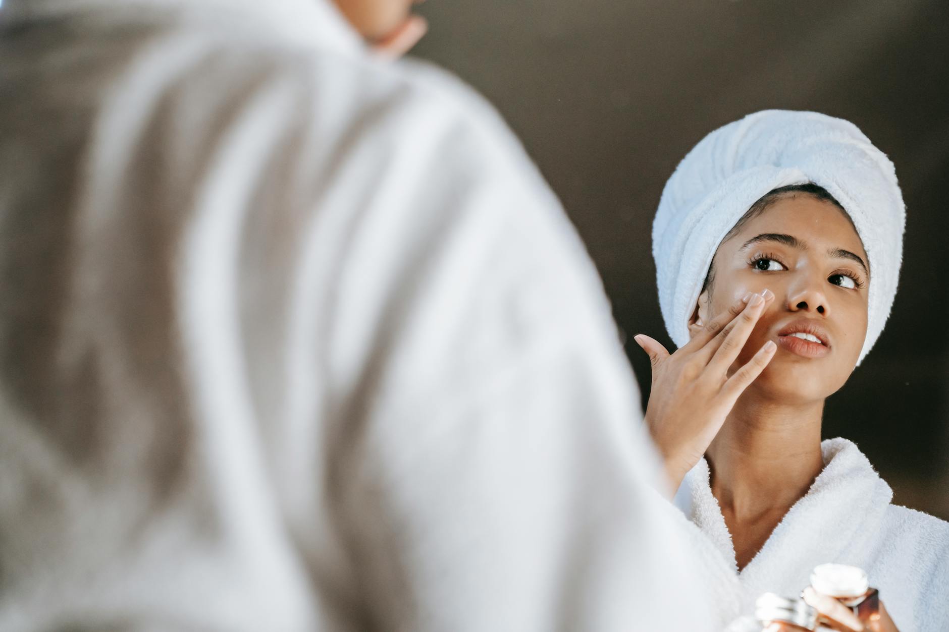 A young woman applies skincare cream while wearing a bathrobe in front of a mirror.Sora Shimazaki