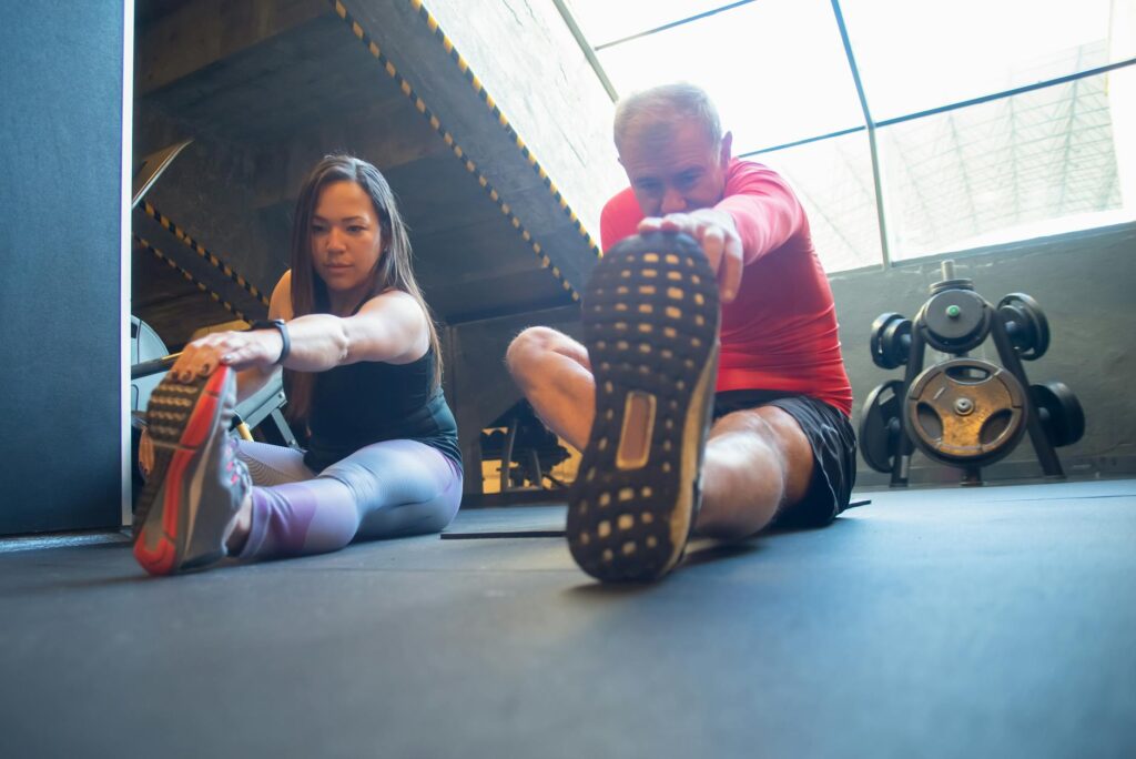 An elderly man and a woman doing stretching exercises on the floor of a gym, promoting a healthy lifestyle.