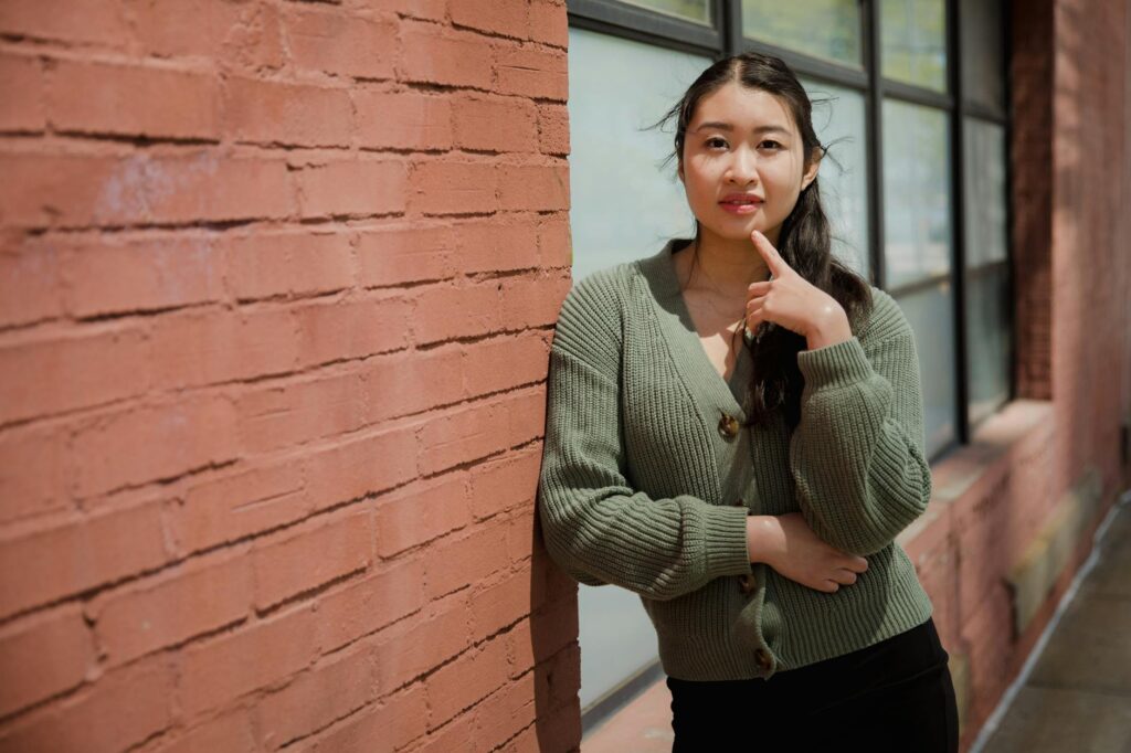 Asian woman in a green cardigan posing against a brick wall during the day.