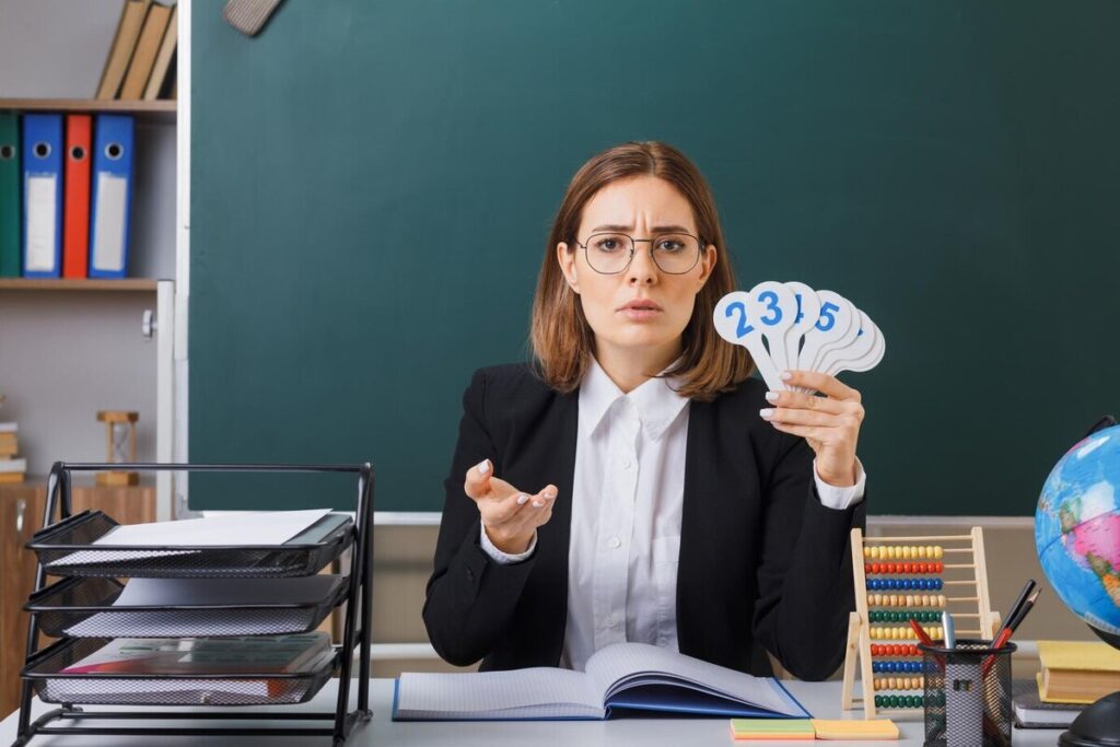 Young woman teacher wearing glasses sitting at school desk in front of blackboard in classroom holding number plates explaining lesson looking displeased raising arm in displeasure