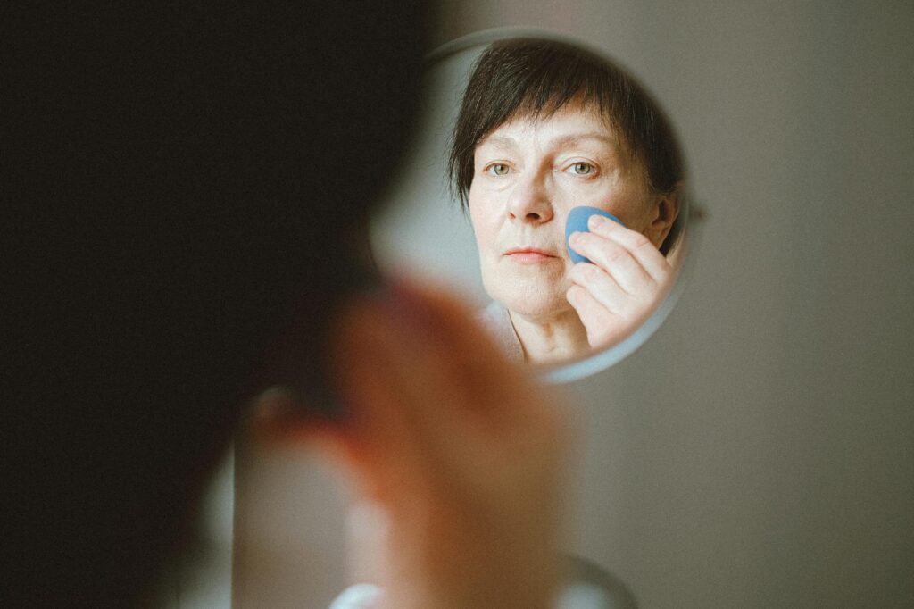 Elderly woman looking at herself in the mirror while applying makeup sponge.Anna Shvets
