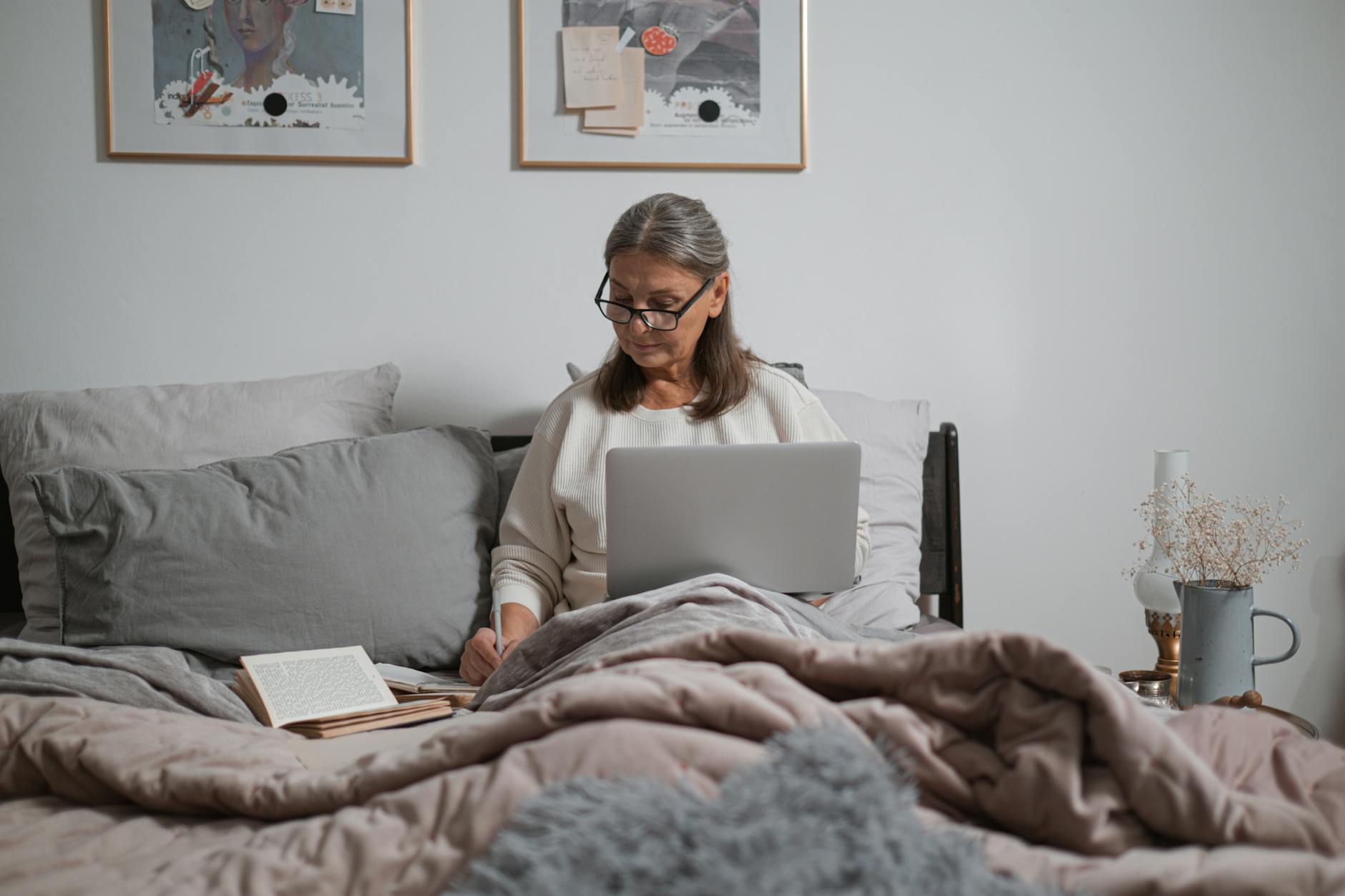 Elderly woman sitting on bed using laptop, writing notes. Comfortable home setting.