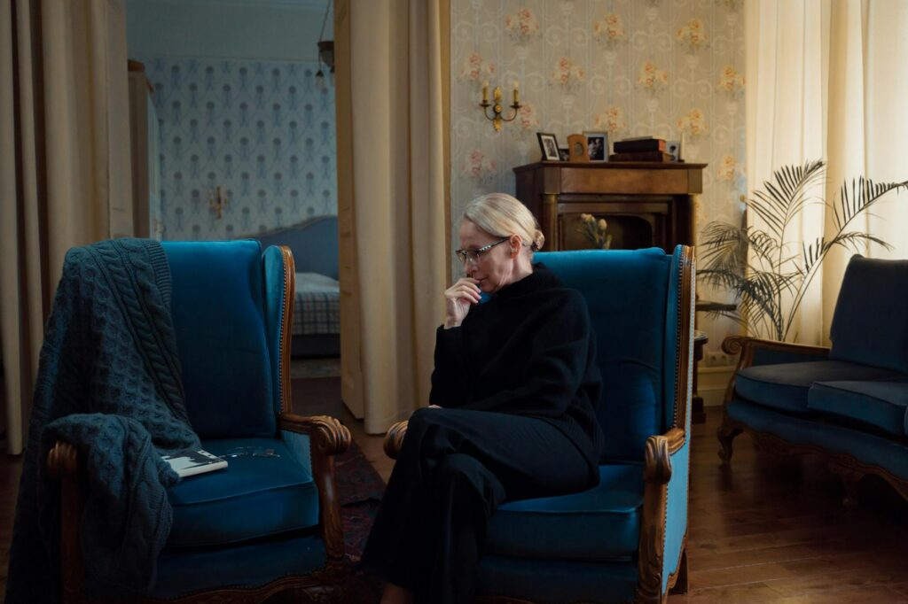 Elderly woman sitting pensively in a cozy living room with vintage decor.