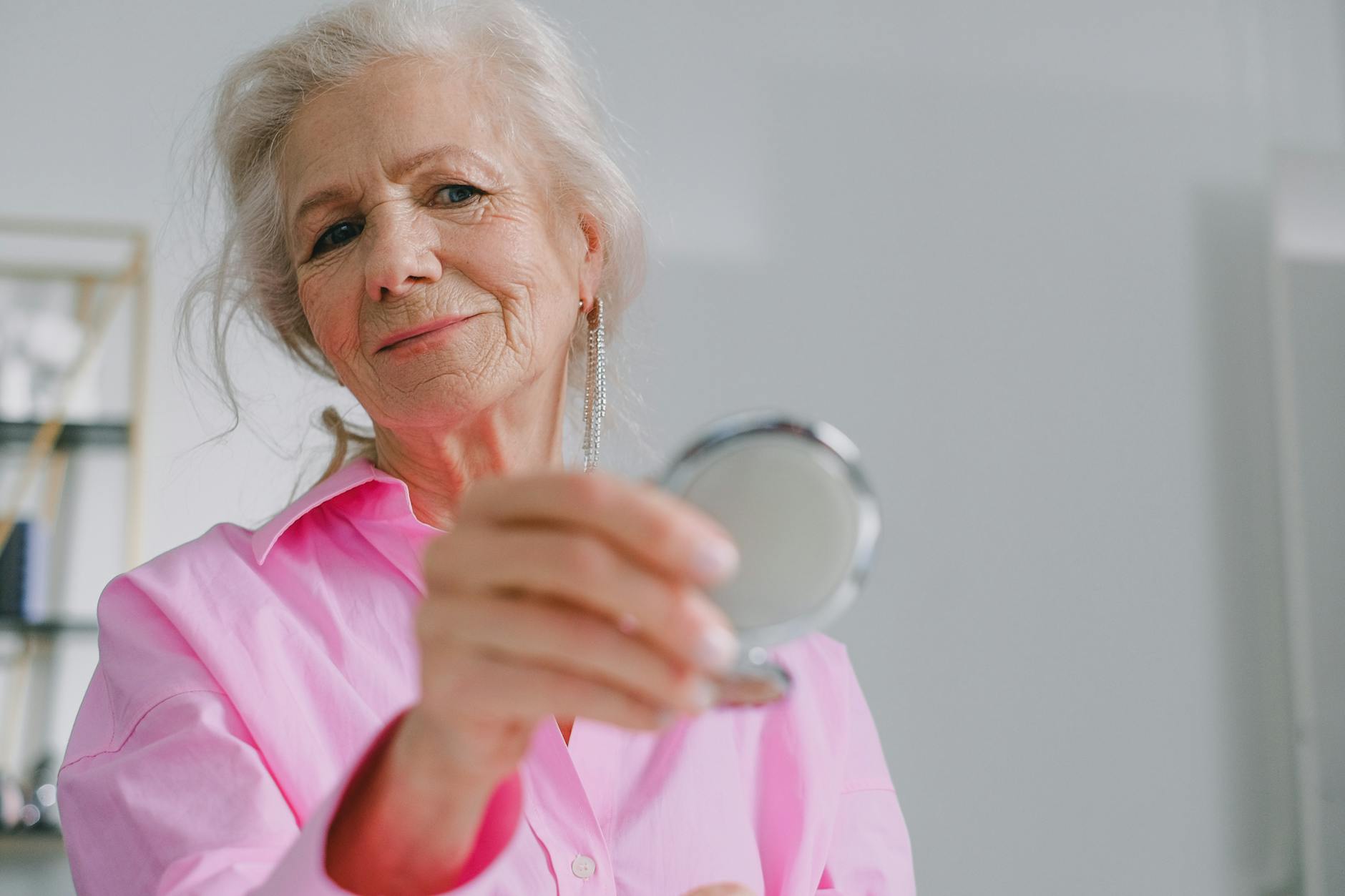 Elegant elderly woman in pink shirt holding a compact mirror indoors reflecting joy and confidence.SHVETS production