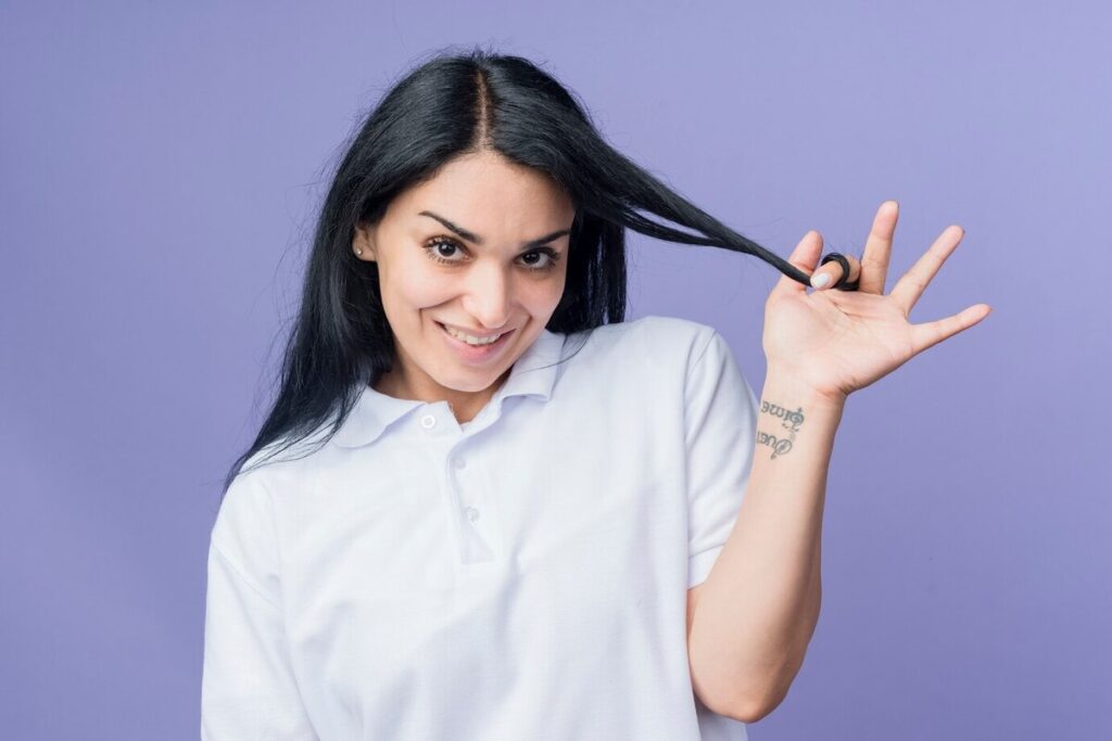 Smiling young brunette caucasian girl rolls hair around finger looking isolated on purple wall