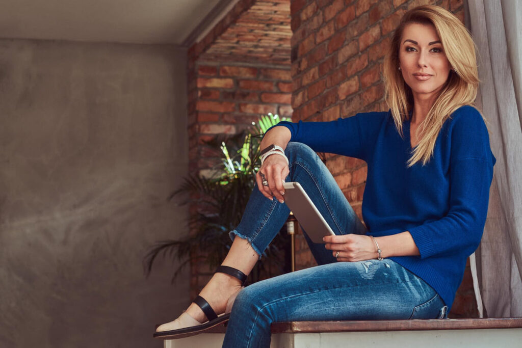 Beautiful blonde female blogger holds a tablet while sitting on a table against a brick wall in a studio with a loft interior.