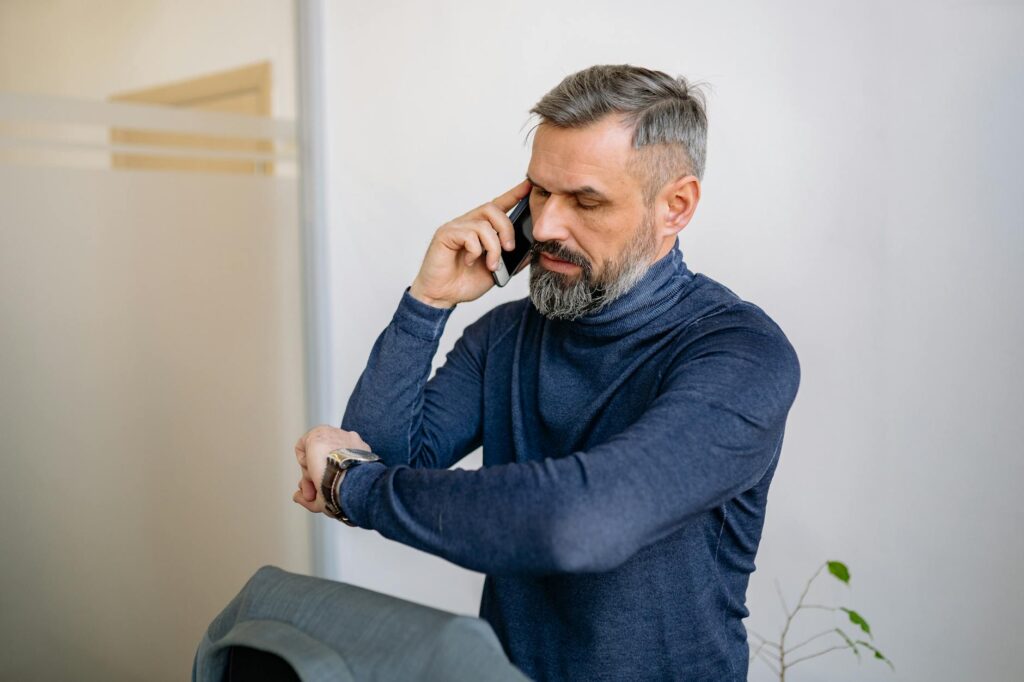 Man with beard speaking on phone while checking the time indoors.Yan Krukau