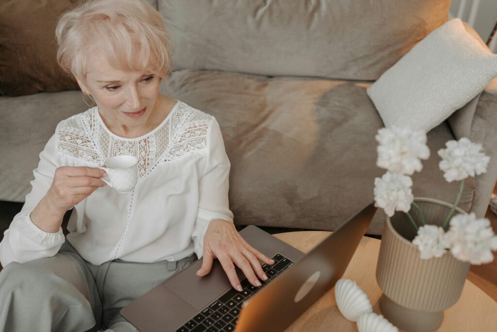 Senior woman enjoying coffee and working remotely from home with a laptop, embracing digital life.