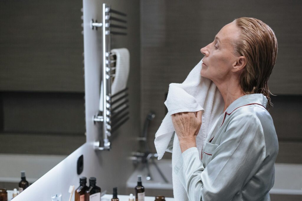 Senior woman in a bathroom, drying her face with a towel during her morning skincare routine.