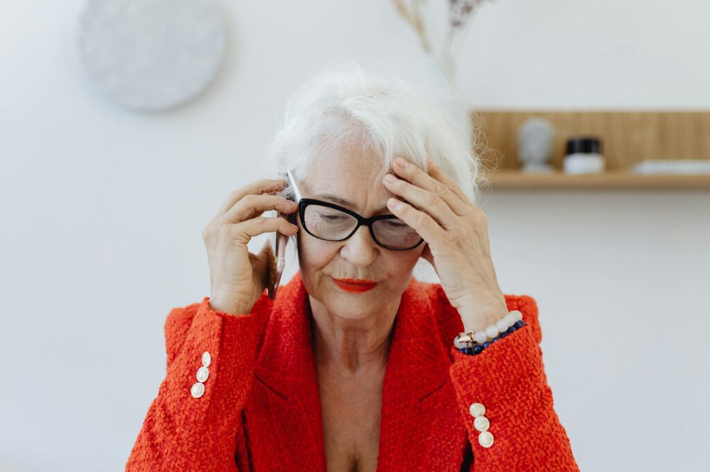 Senior woman in red blazer stressed while talking on the phone indoors.Photo By Kaboompics.com