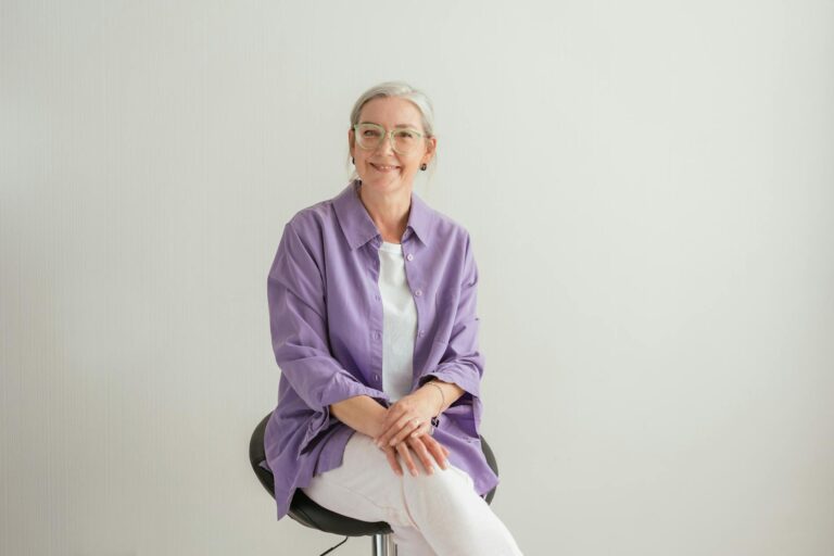 Senior woman with eyeglasses smiling in casual purple blouse, seated indoors.
