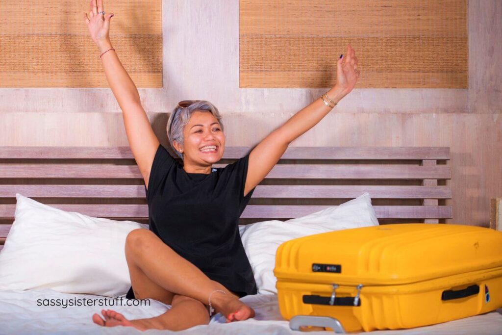 Asian woman sitting on a hotel bed with her large suitcase next to her and her hands in the air with excitement.