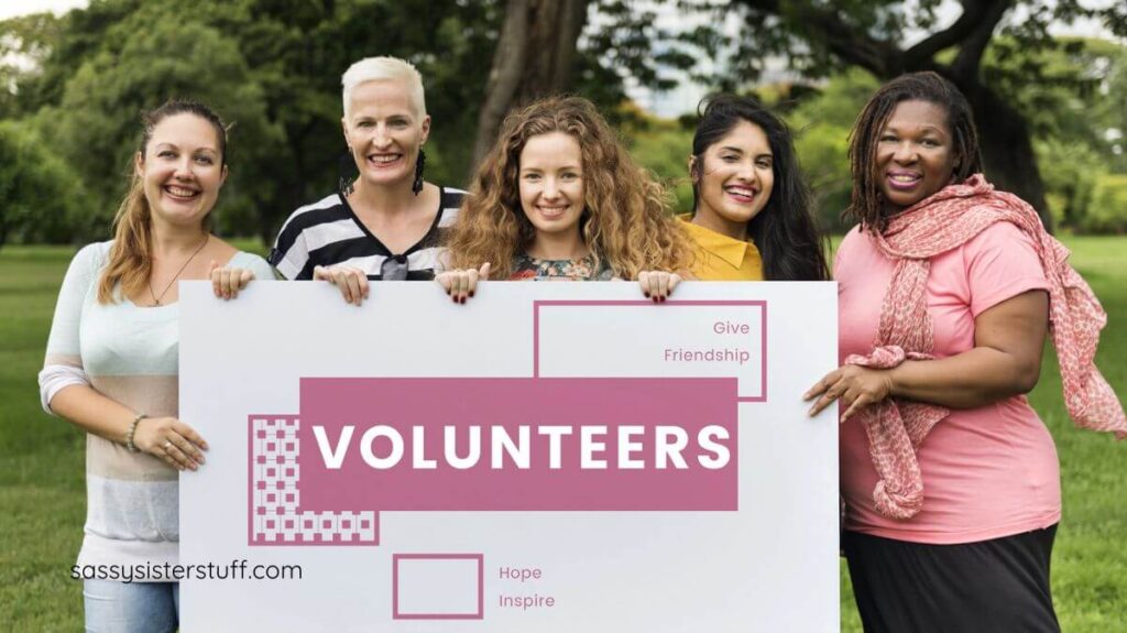 five middle aged women holding a large sign that says VOLUNTEERS.
