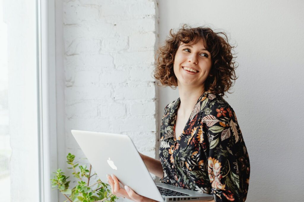 Woman with curly hair holding a laptop while smiling near a window indoors.