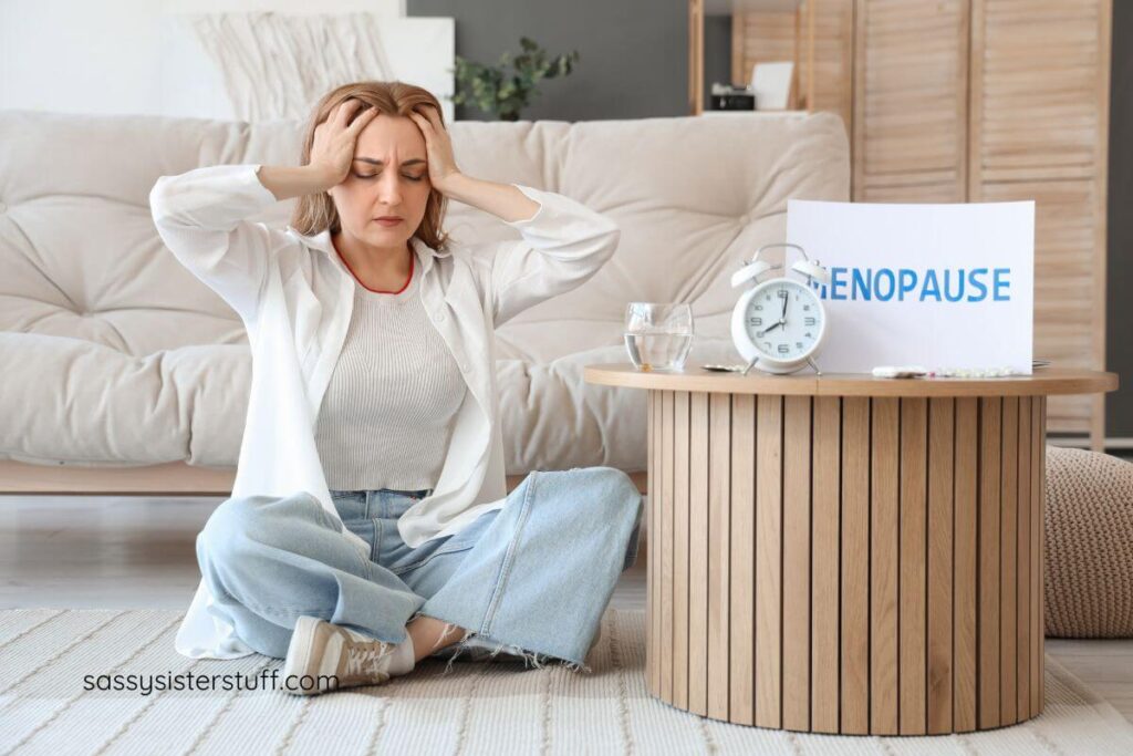 midlife woman holds her head in both hands, sitting on the floor, with a sign next to her that says menopause.