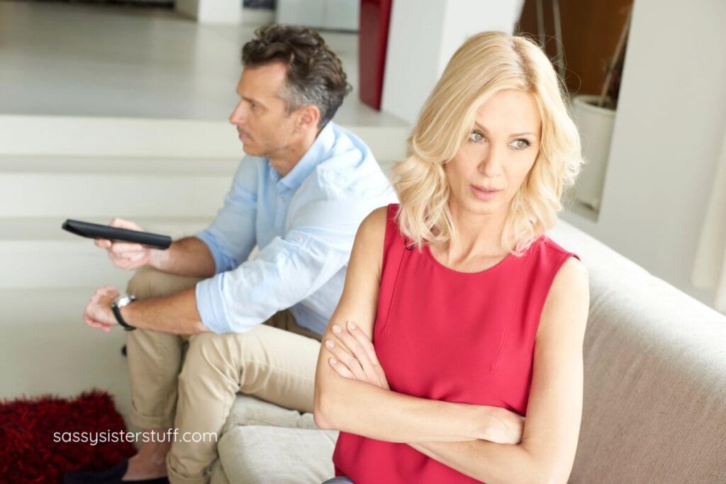 midlife woman is frustrated with her husband who is watching tv in the background.