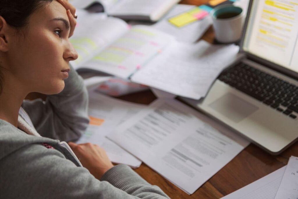 woman sits at desk filled with paperwork and has her head resting on her hand in frustation and overwhelm.