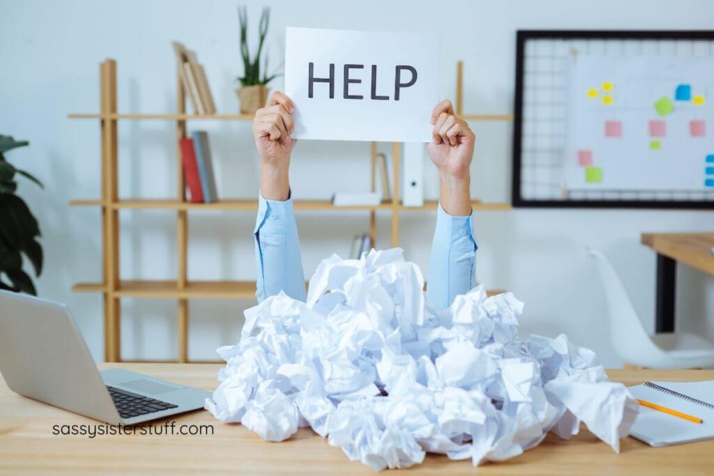 worker at a laptop holds a HELP sign up from behind a big pile of crumbled up paper to demonstratre ways for independent people to practice asking for help.