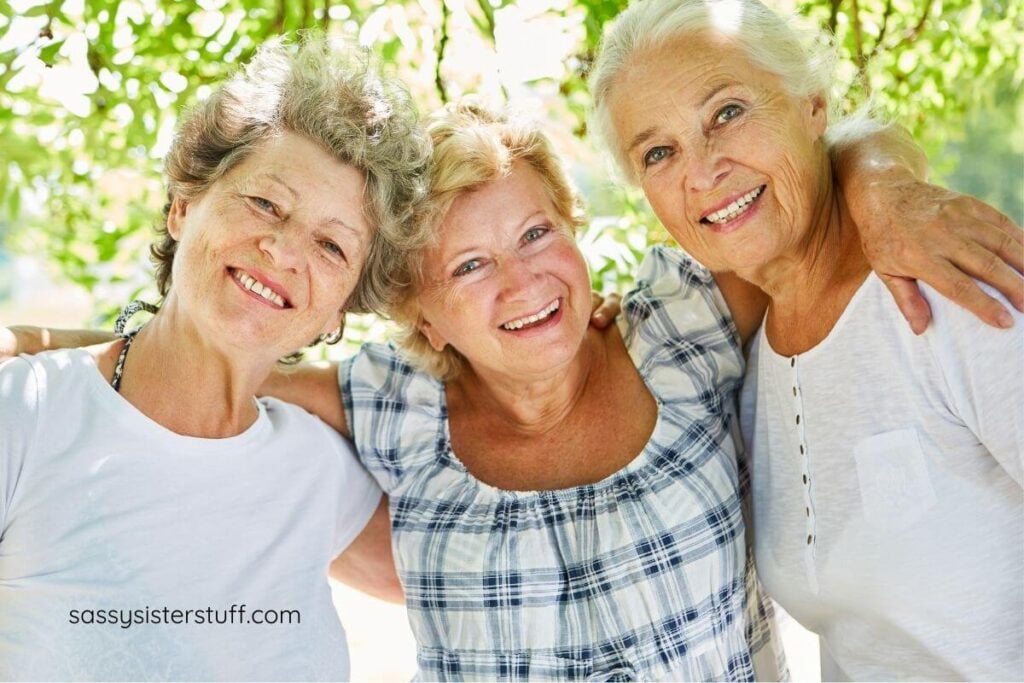 three mature women dressed casually in light colored blouses hugging and smiling at the camera.