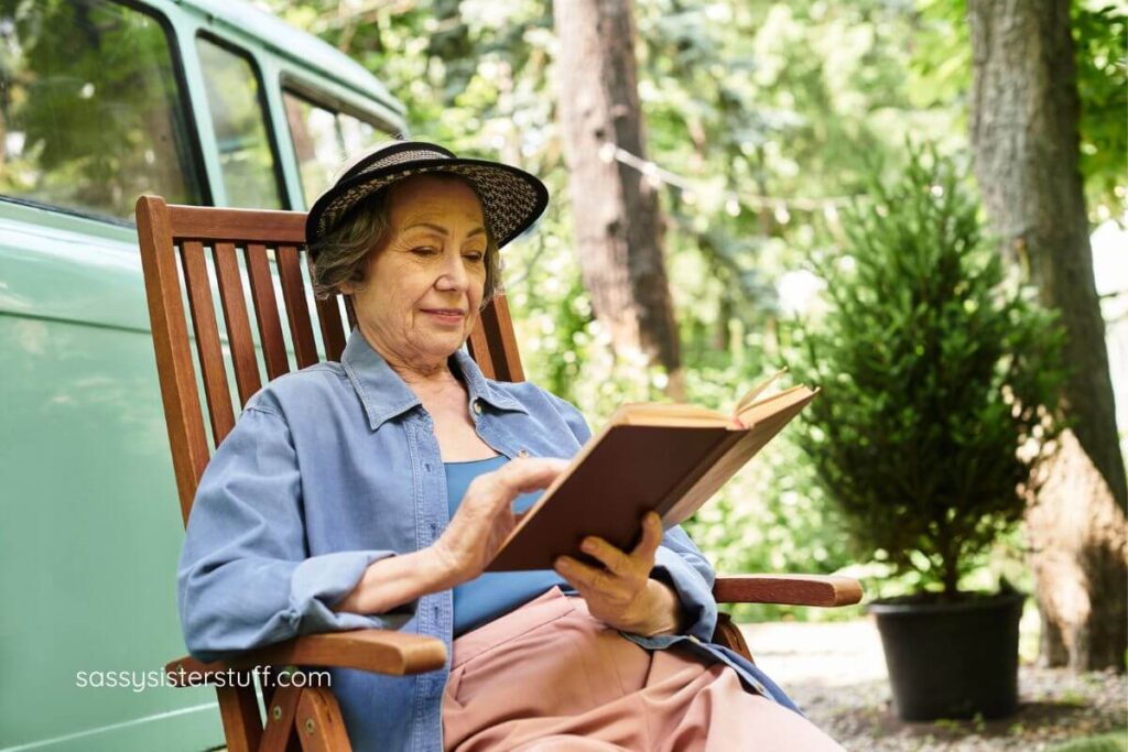 senior woman sits in a wooden chair outside in a campground and relaxes while reading a book.
