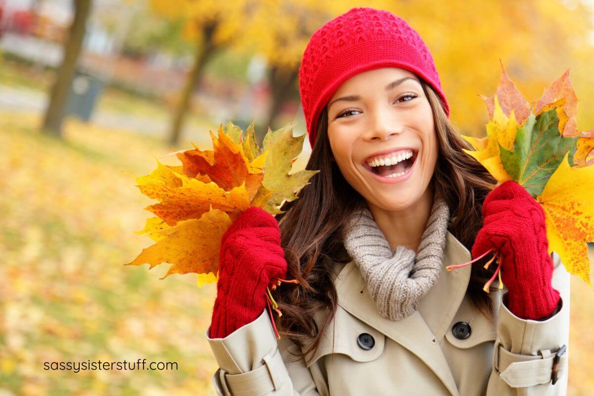 women dressed for Autumn has gathered colorful leaves and has them in her hands, smiling real big in the park.