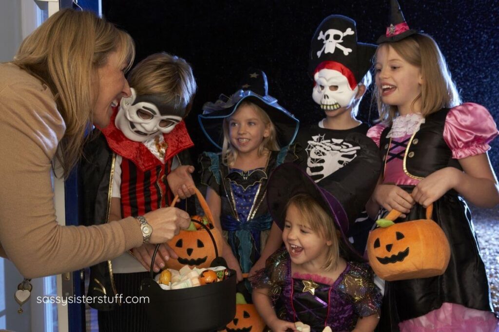five excited children trick or treat at a house where a middle aged woman has lots of treats for them.
