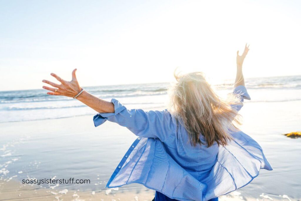 happy midlife woman throwing her hands up in the air on the shore of the ocean to celebrate the changes that come with age.