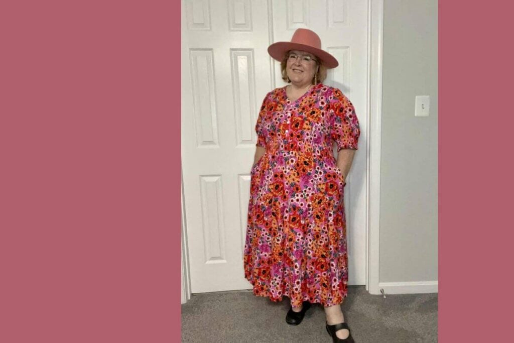 sassy sister stuff founder models a floral dress in shades of pinks, reds, and corals with a coral hat.