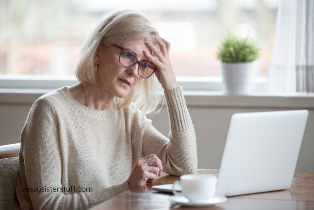 middle-aged woman sitting at a table with a laptop has her hand up to her forhead because she can't focus and get out of her own way.