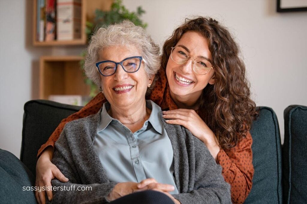 adult daughter hugs her elderly mother around her shoulders and they smile at the camera.
