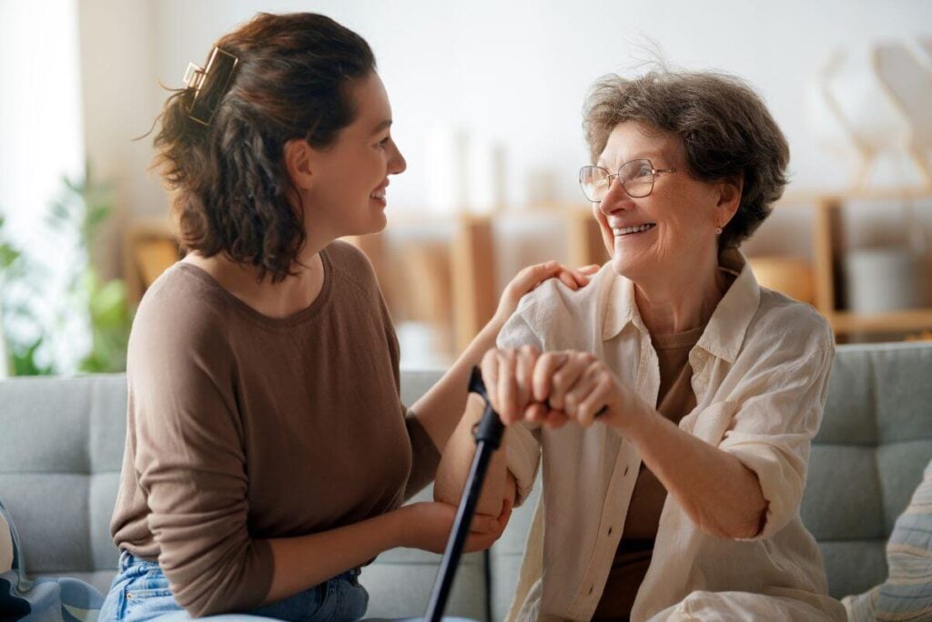 adult daughter sits on a sofa with her elderly mother who is holding a cane.