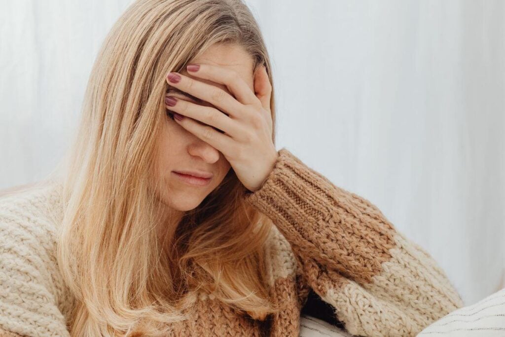 adult daughter sits on a sofa with her hand covering her eyes feeling exhausted.