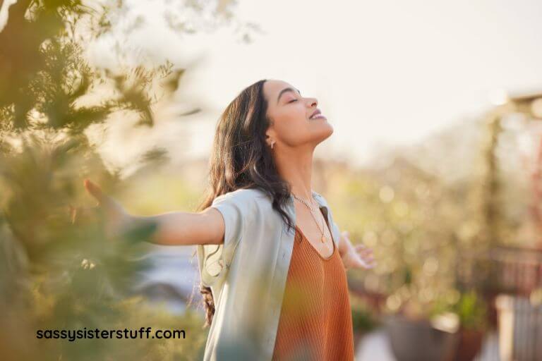 young woman stands outside in the sun with her face looking up, her eyes closed, and her arms stretched out meditating.