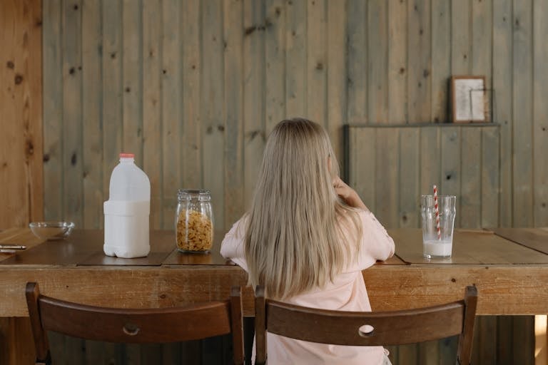 A young girl with long hair sits at a wooden dining table, enjoying breakfast with milk and cereal.
