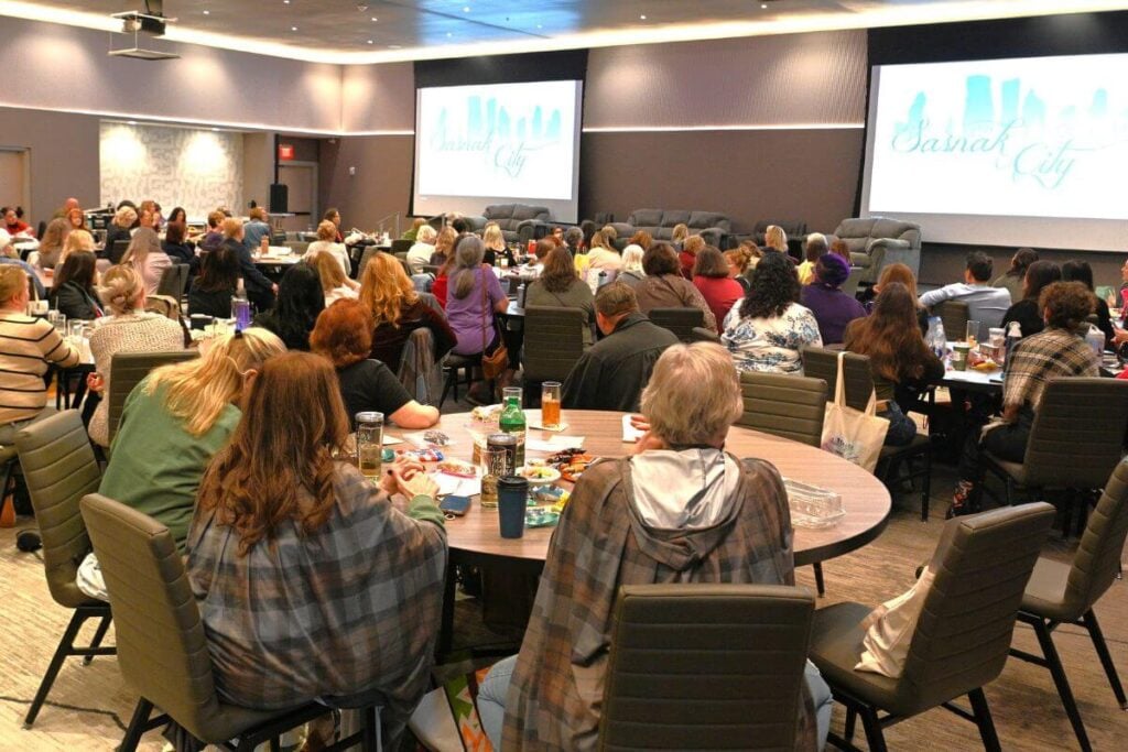 a large group of women sit at round tables in an auditorium listening to a presentation.