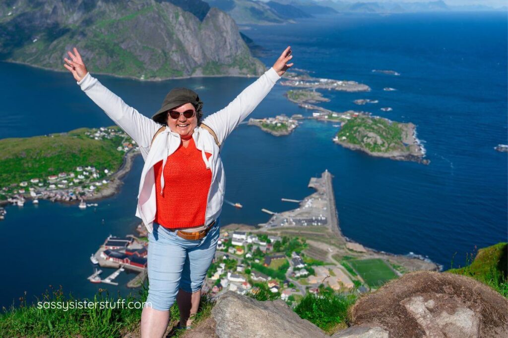 middle aged woman with arms outreached stands on top of a mountain with water and islands in the background.